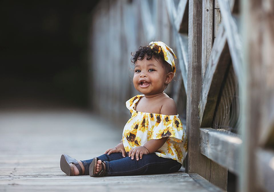 Adorable toddler girl in yellow sunflower outfit sitting on a wooden bridge walkway. Midlothian VA Photographer