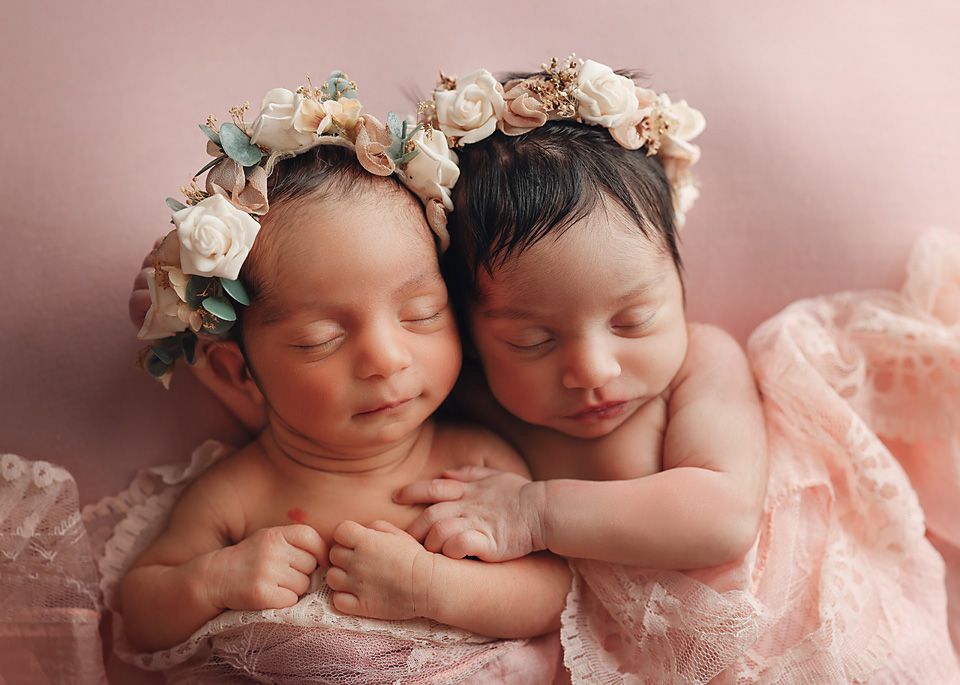 Two sleeping newborn twin babies wearing floral crowns cuddle together on a soft pink backdrop.