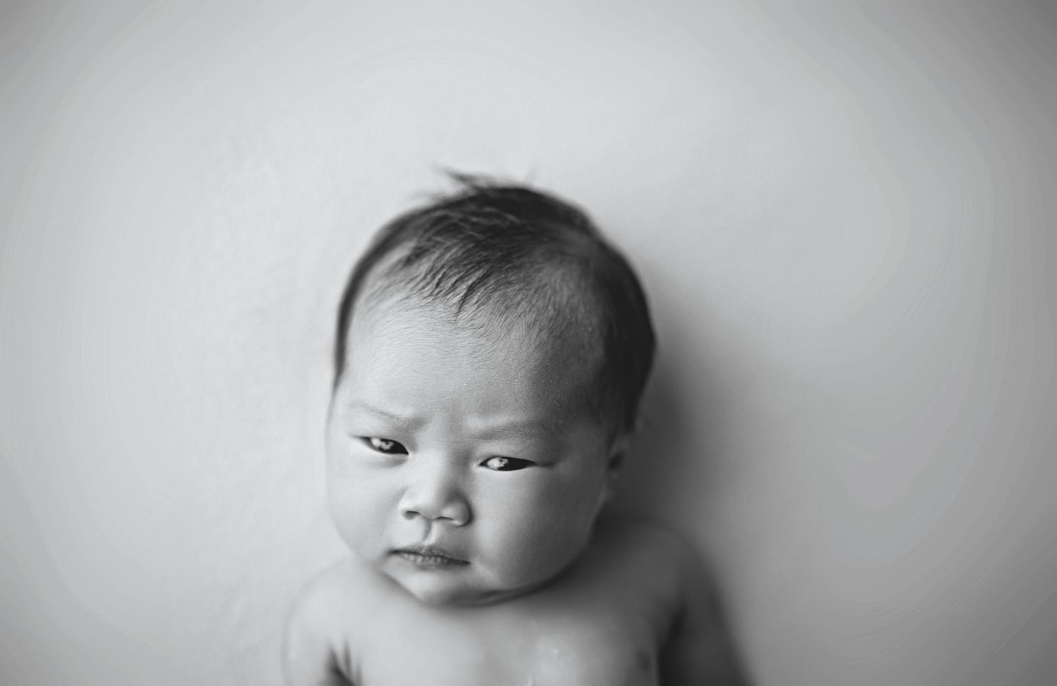 Black and white newborn baby portrait with dark hair, looking downward with a serious expression.