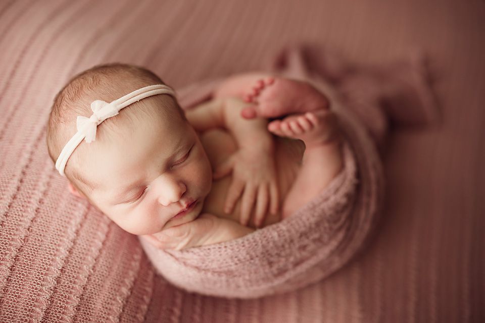 Sleeping newborn baby girl wrapped in pink with a bow headband curled in a round pose.