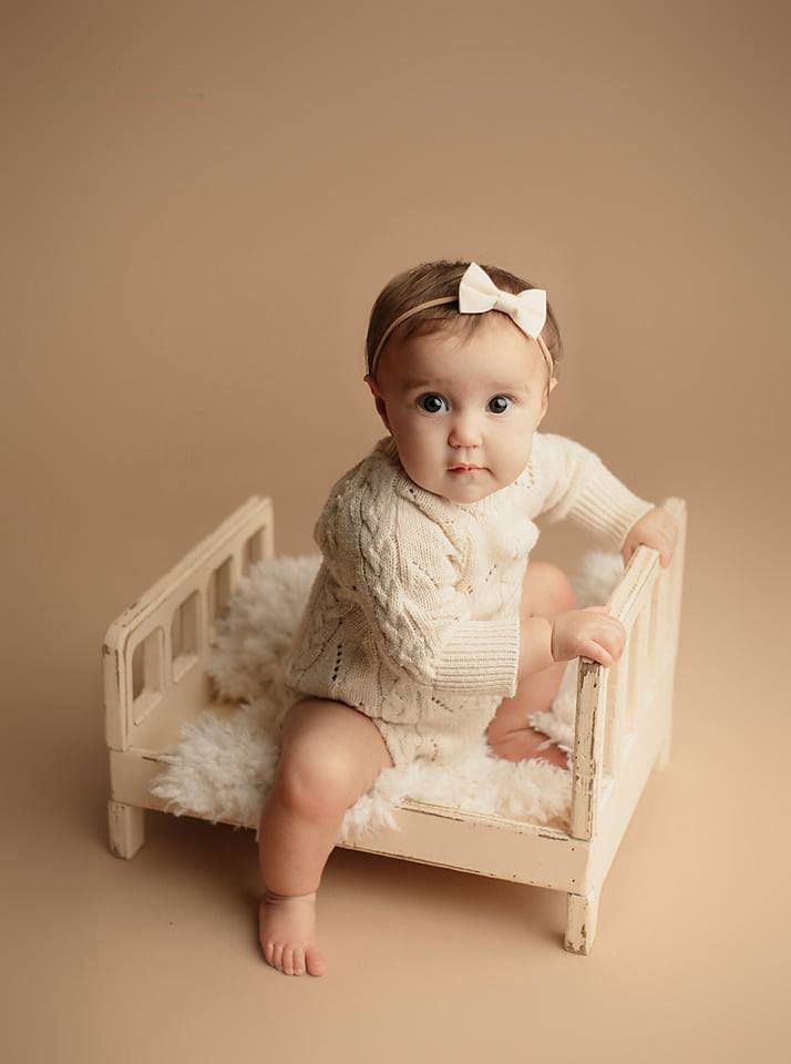 Adorable baby girl in cream knit romper and bow headband standing beside a small vintage white bed.
Chesterfield va photographer in studio