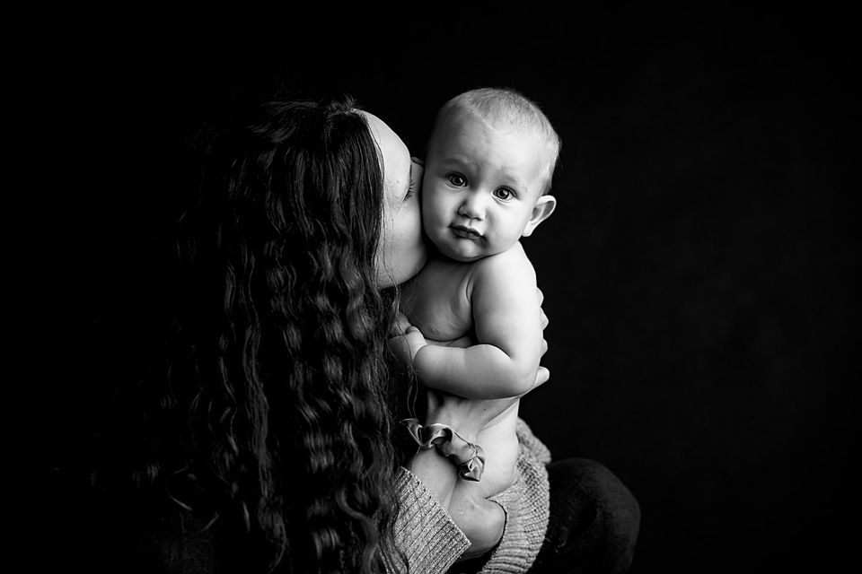 Black and white photo of a mother kissing her baby on the cheek against a dark background.