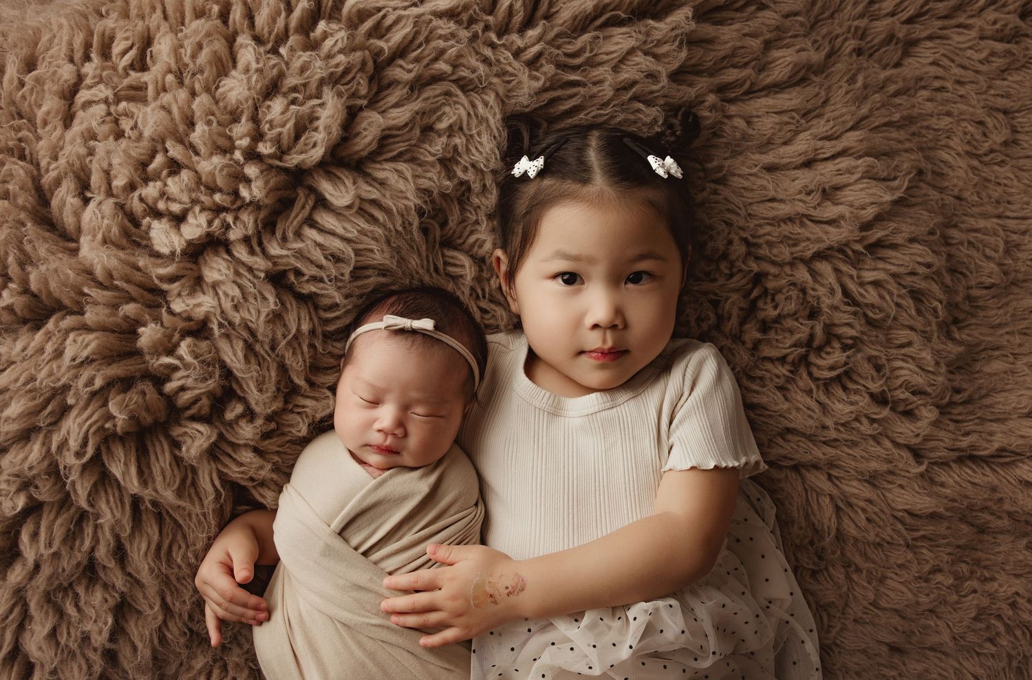 Older toddler girl with hair clips holds sleeping swaddled newborn baby on fluffy brown rug.