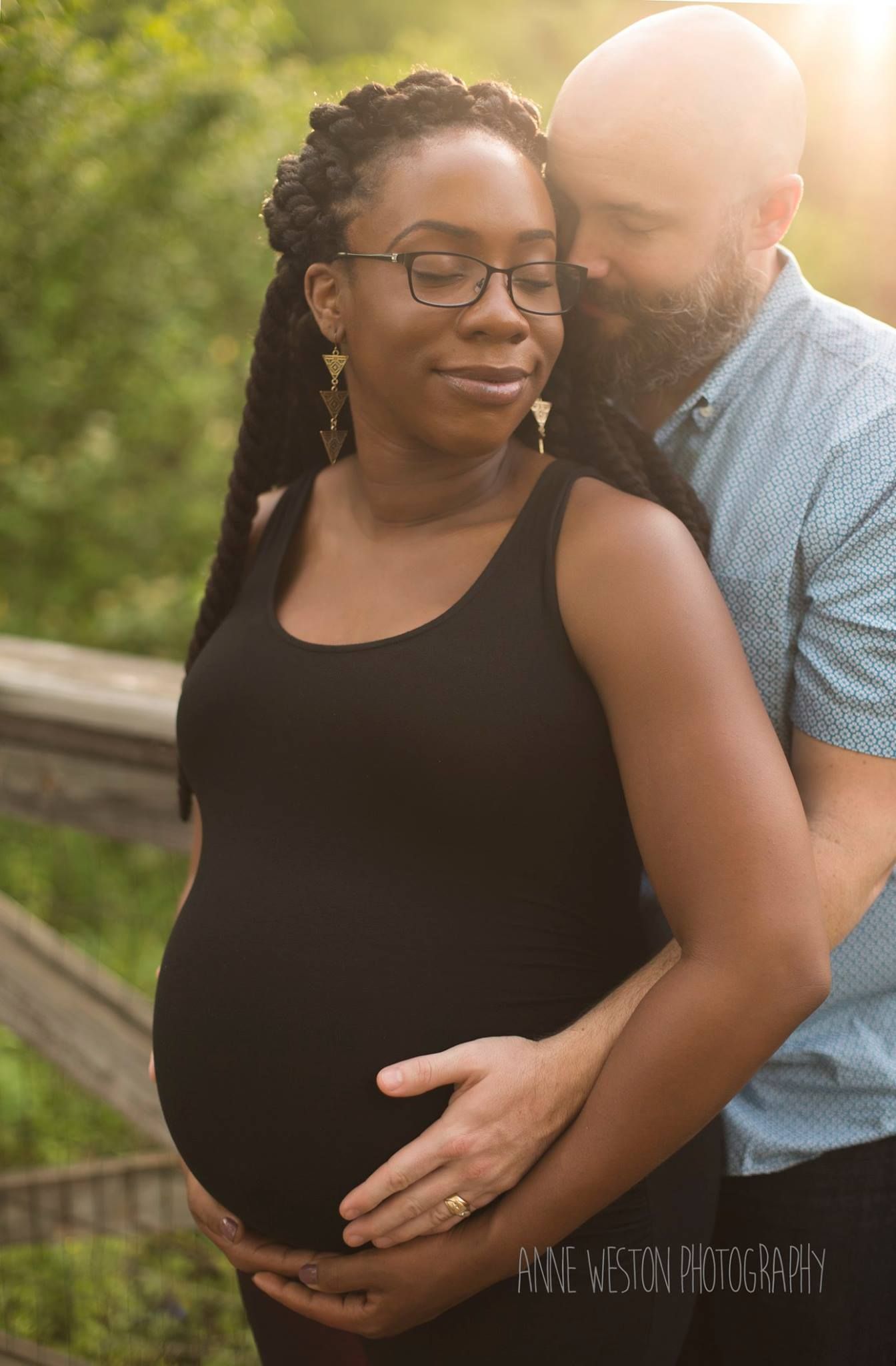 Pregnant woman in black dress embraced by bearded man outdoors in golden hour light, maternity photography session. Midlothian, VA