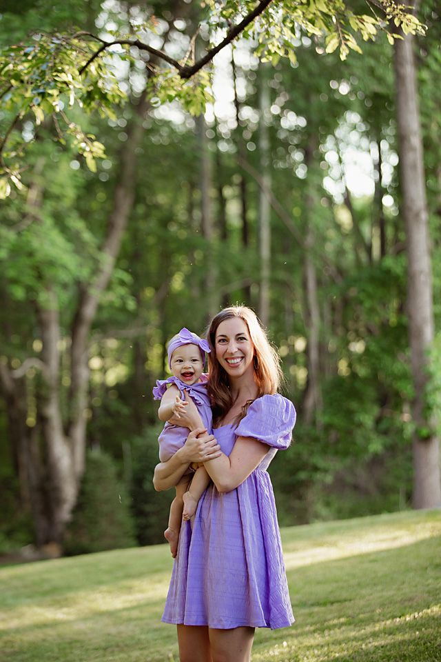 Mother in lavender dress holds her laughing baby girl in matching purple outfit in a sunny green park.