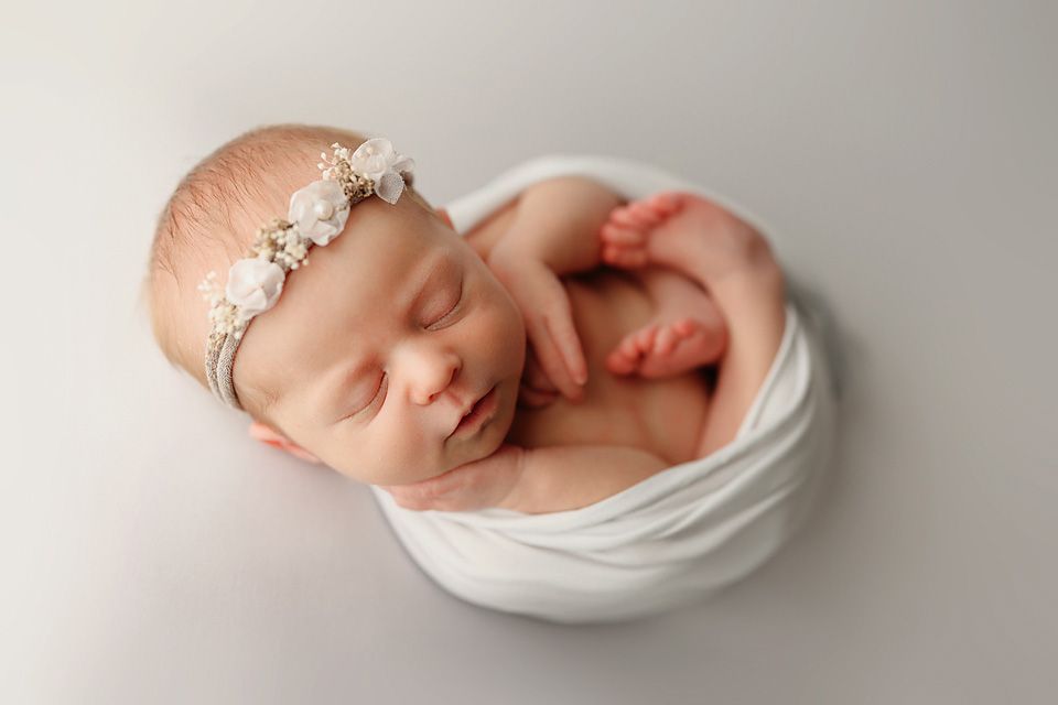 Sleeping newborn baby wrapped snugly in white cloth with decorative headband on gray background.