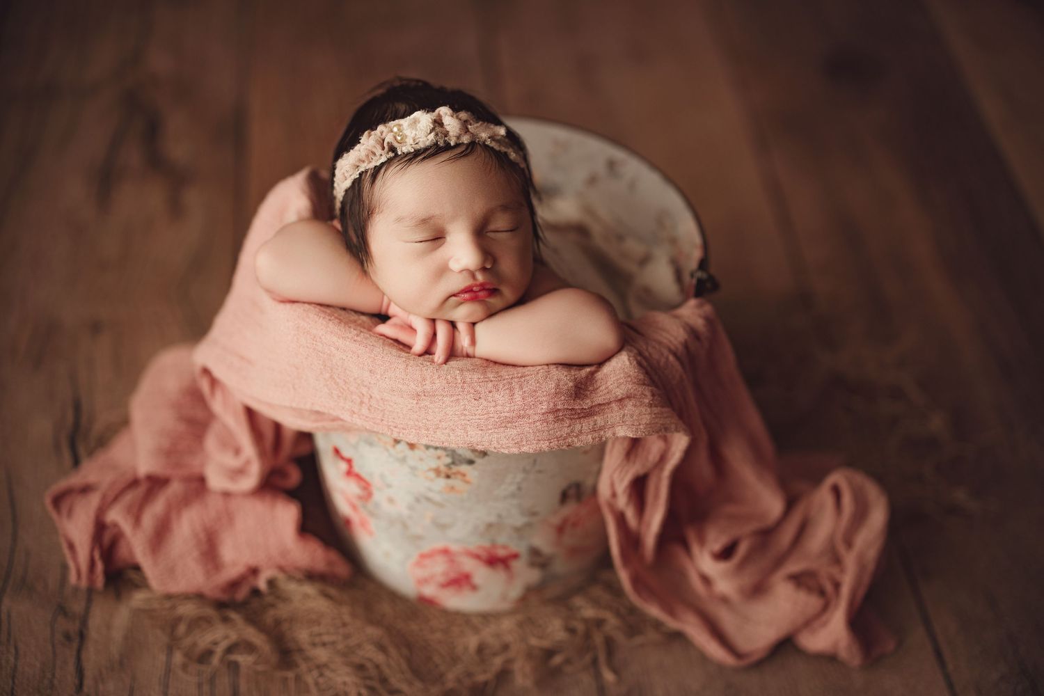 Sleeping newborn baby girl in floral bucket with pink wrap and headband on wooden floor.