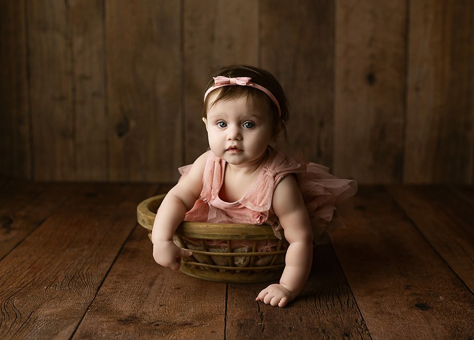 Baby girl in peach tutu dress sitting in a wicker basket on a rustic wooden floor.