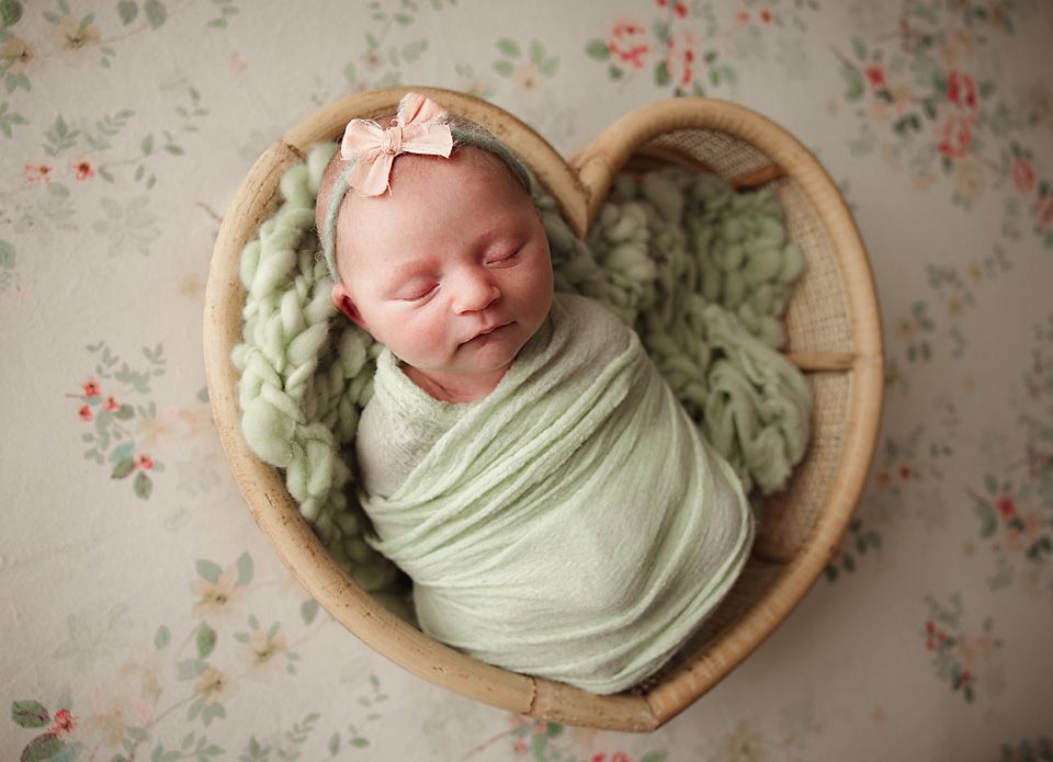 Sleeping newborn baby wrapped in green swaddle with pink bow headband in heart-shaped wicker basket.