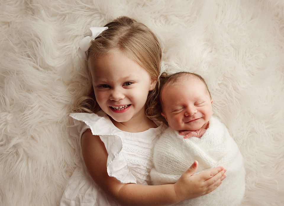 Smiling toddler girl in white dress hugging sleeping swaddled newborn sibling on fluffy white rug.
