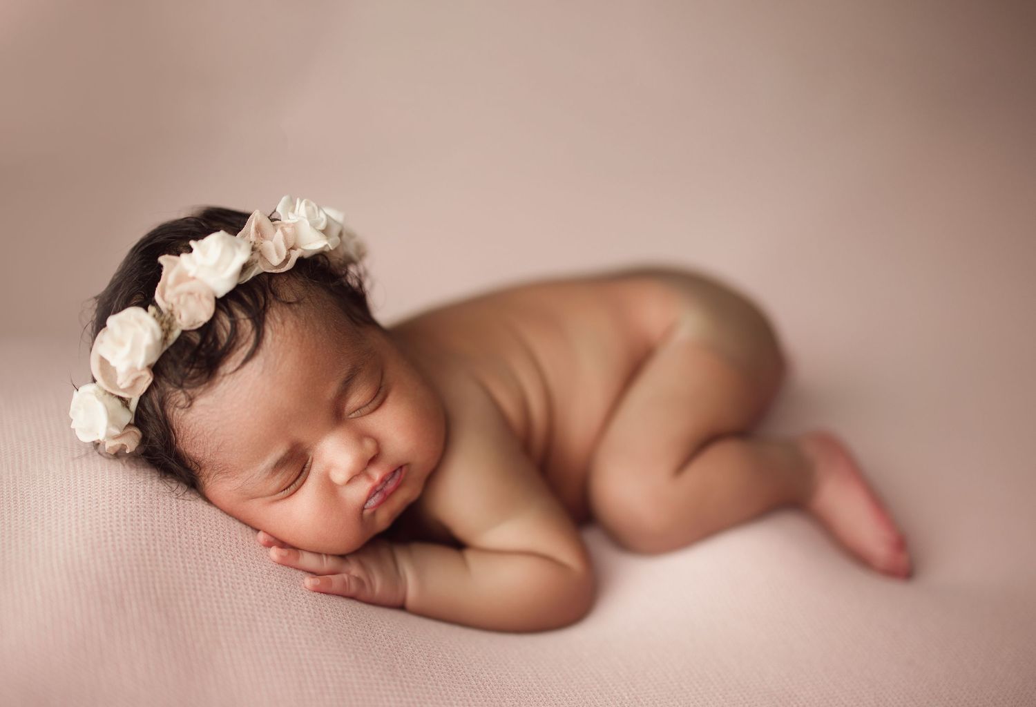Sleeping newborn baby girl wearing a delicate white floral crown, posed on a soft pink blanket.