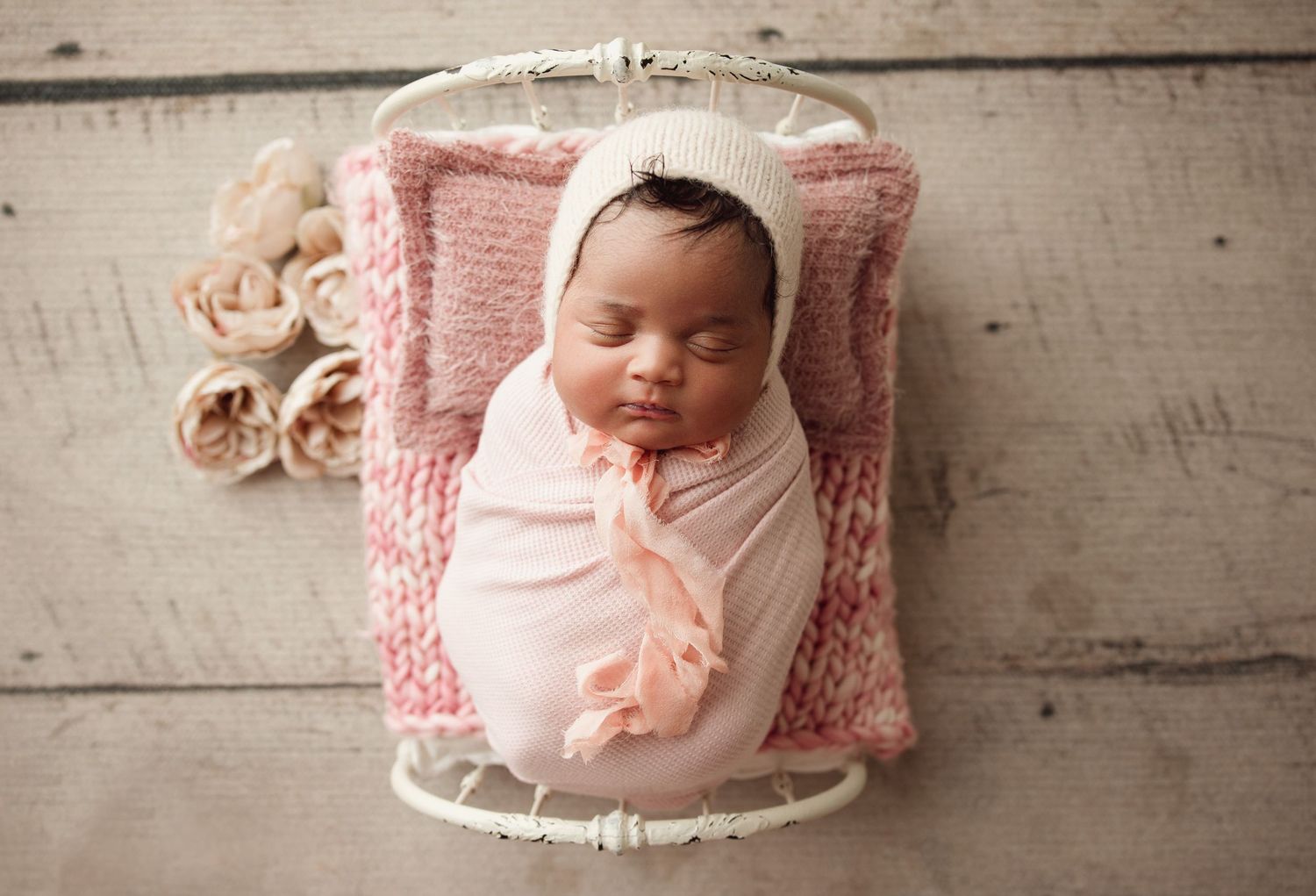Newborn baby swaddled in peach wrap, sleeping peacefully in a white mini crib with pink knit blanket and roses.