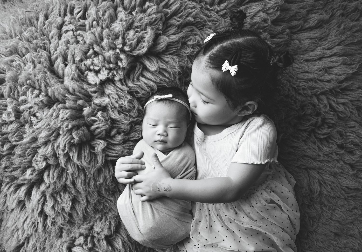 Black and white photo of a young girl tenderly holding a swaddled newborn baby sibling on a fluffy rug.