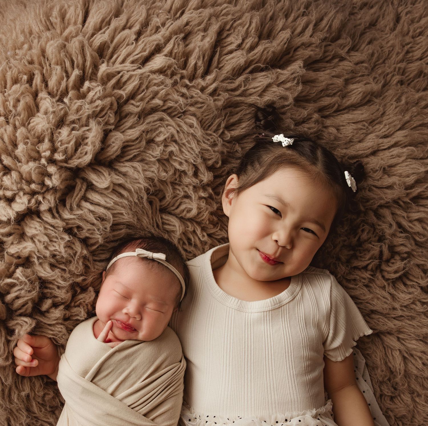 Newborn baby swaddled beside smiling toddler girl with pigtails on fluffy brown rug.