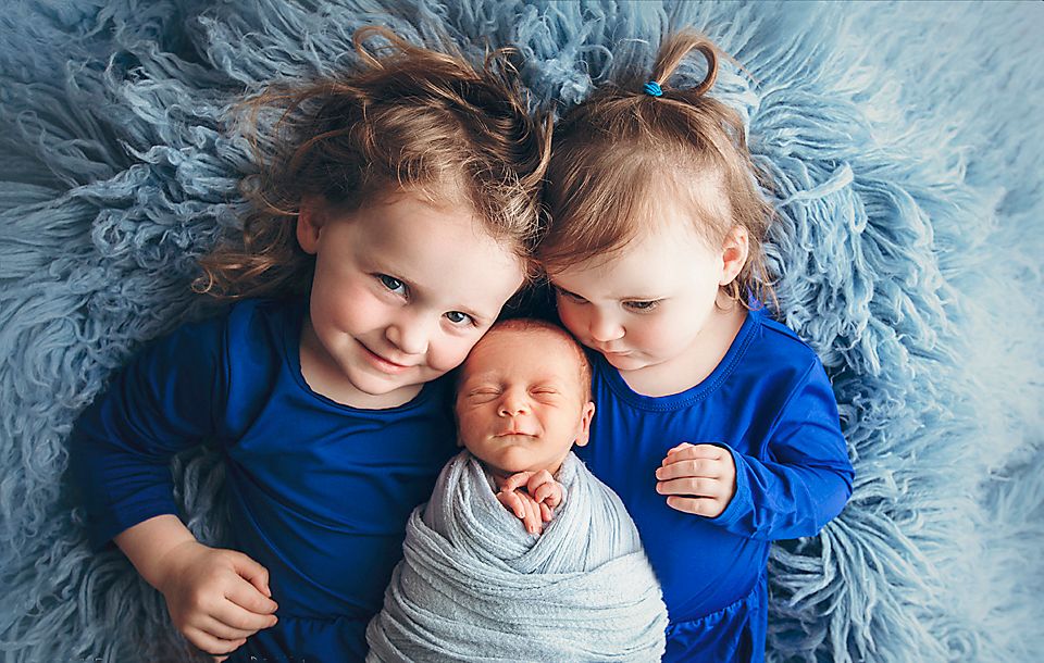 Two young siblings in blue outfits cuddling their swaddled newborn baby sibling.
