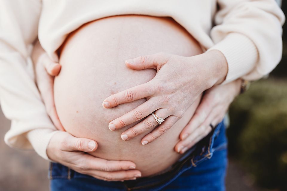 Close-up of pregnant woman's baby bump with hands gently cradling it, wearing a ring and white top.Richmond newborn photographer