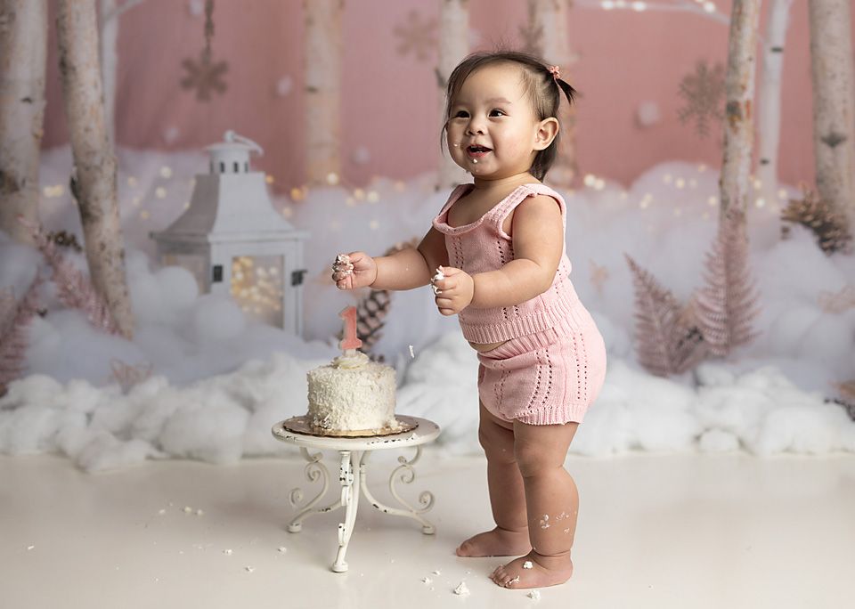 Happy baby girl in pink romper smashing a white cake in a winter wonderland studio setting.