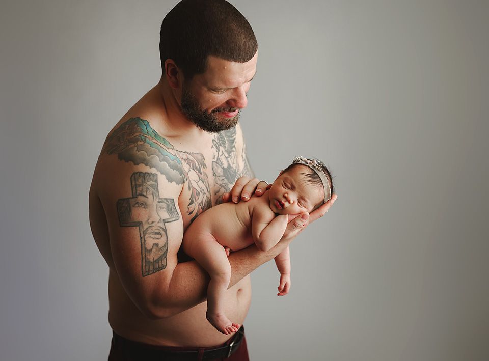 Tattooed shirtless father tenderly holds sleeping newborn baby girl wearing headband against gray background Richmond newborn photographer