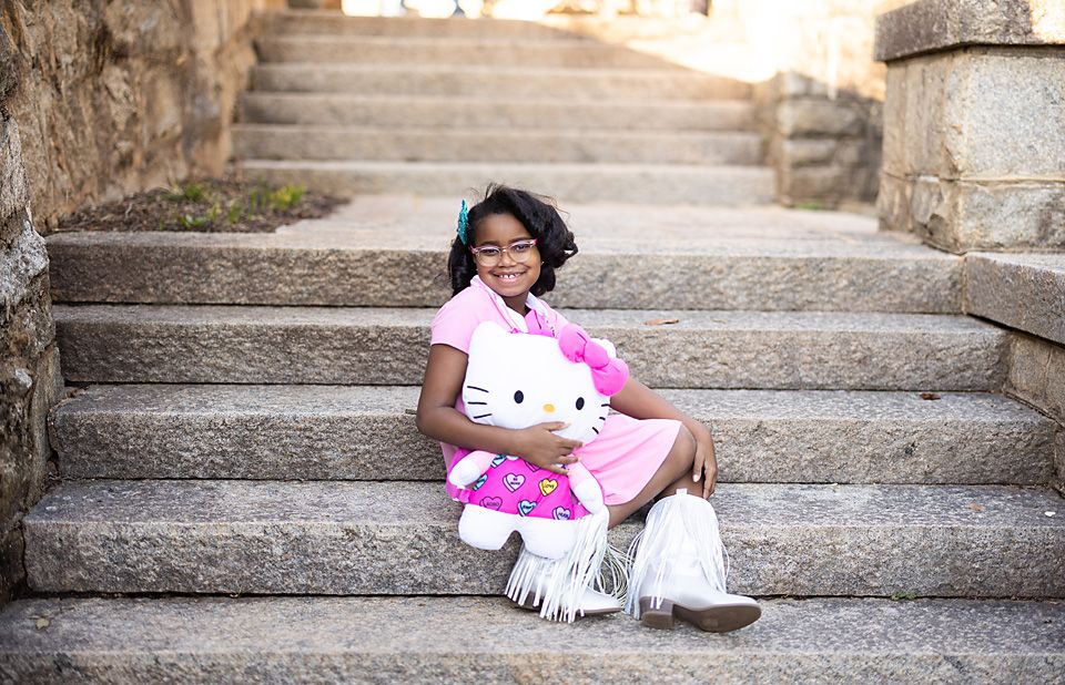 Young girl in pink outfit sitting on stone steps holding a Hello Kitty plush toy.Richmond va family photography.
