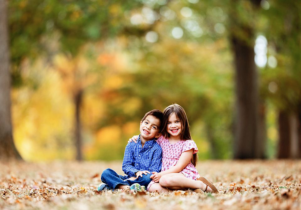 Two young children sitting together in fallen autumn leaves in a park. fall mini family session photographer