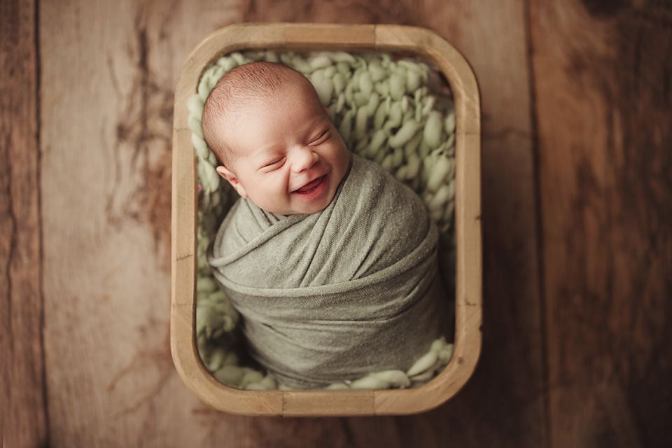 Smiling newborn baby swaddled in green wrap nestled in wooden box with soft green blanket.