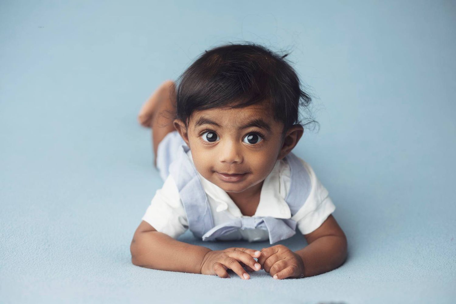 Curious Indian baby boy in white shirt propped on elbows during blue backdrop photo session.