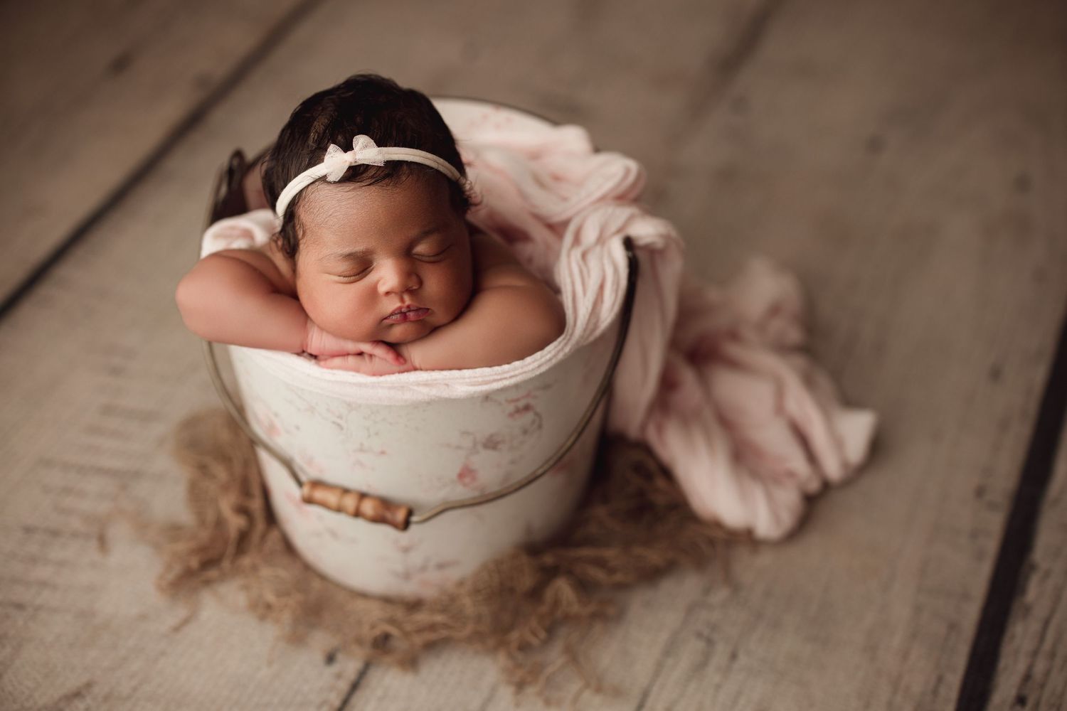 Newborn baby sleeping peacefully in a white bucket with pink wrap and headband on rustic wood floor.