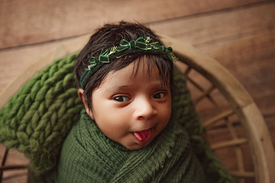 Adorable dark-haired newborn baby wrapped in green swaddle with floral headband sticking tongue out.