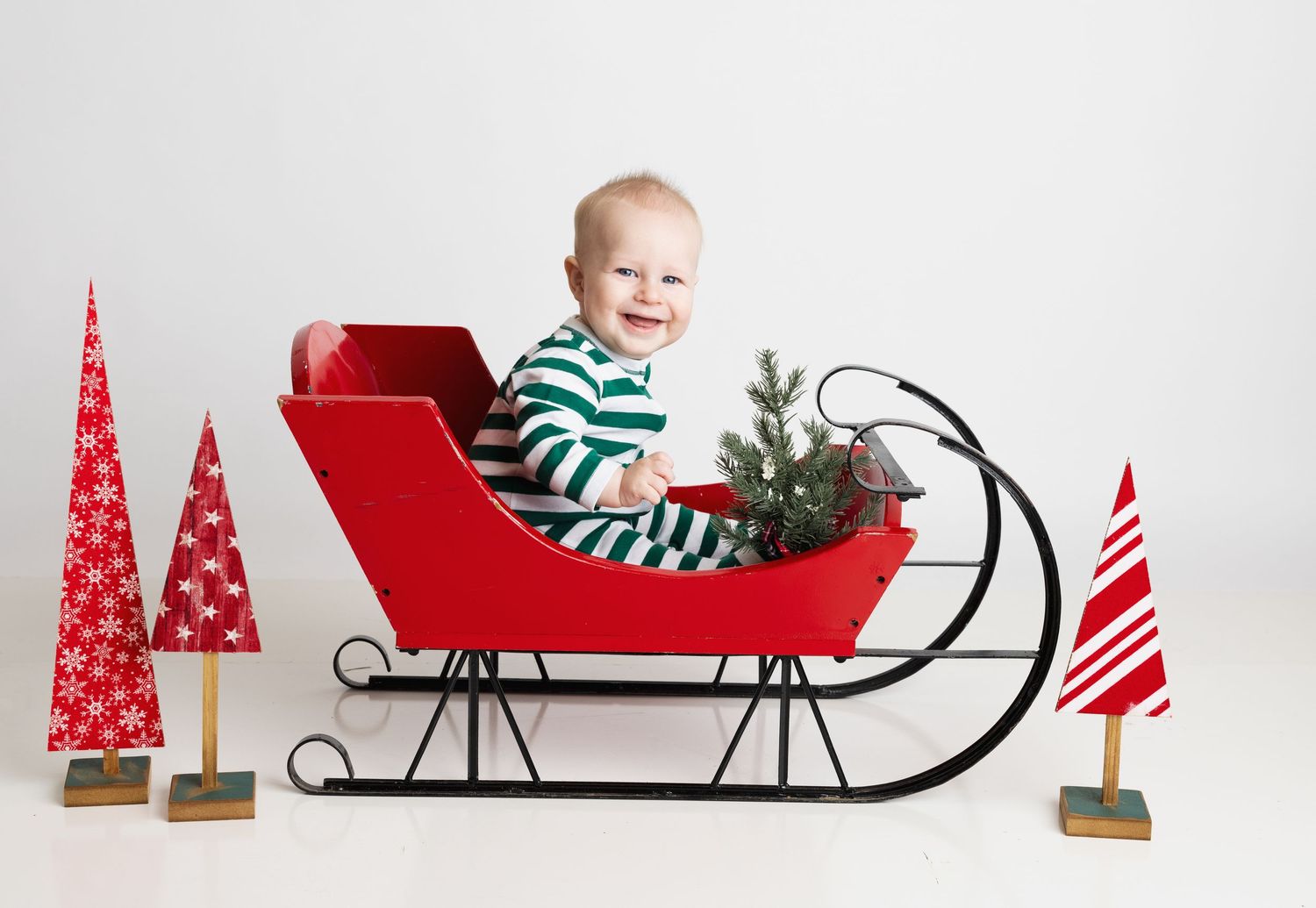 Baby in green striped pajamas sitting in a red Christmas sleigh with festive trees on a white background Richmond newborn photographer