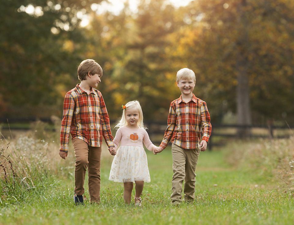 Three siblings walk hand-in-hand through a green field during a fall family photo session.