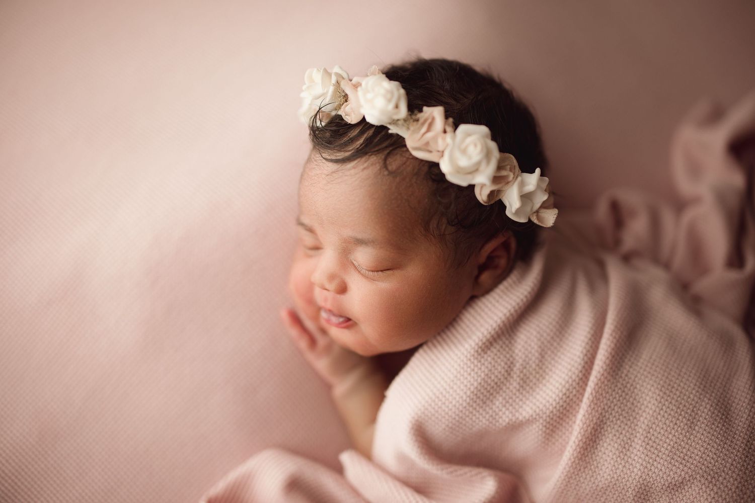 Sleeping newborn baby girl wrapped in pink with a delicate white floral headband on a soft pink background.
