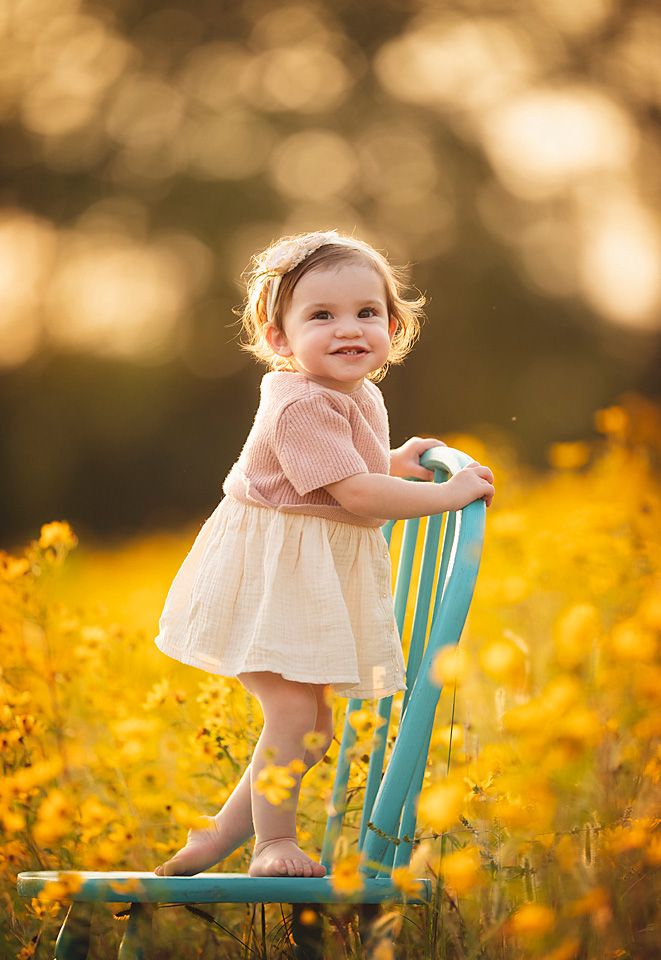 Smiling toddler girl in pink dress standing on teal chair in golden sunlit wildflower field.