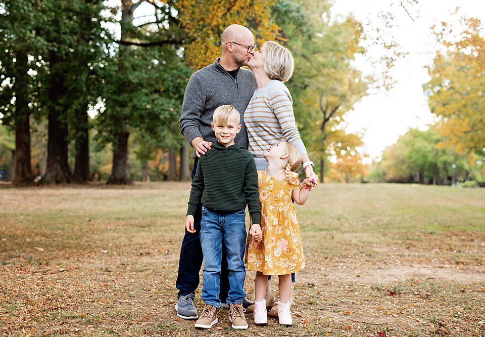Family of four posing in an autumn park, parents kissing while children stand nearby.