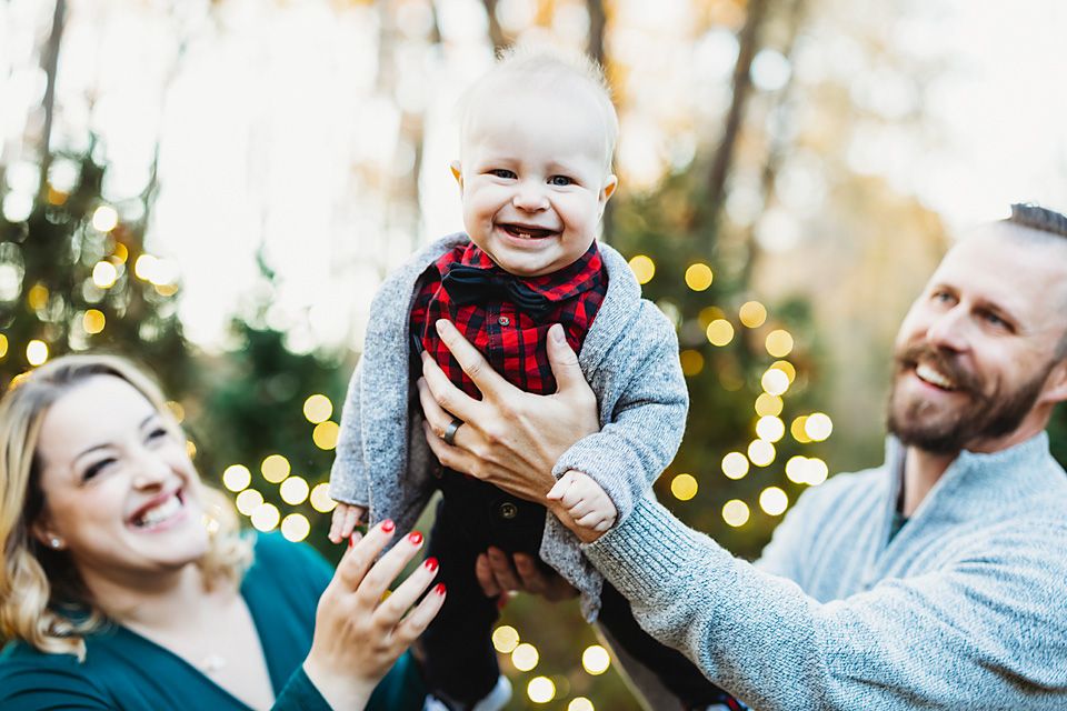 Joyful family holiday portrait with parents lifting laughing baby boy near Christmas lights.