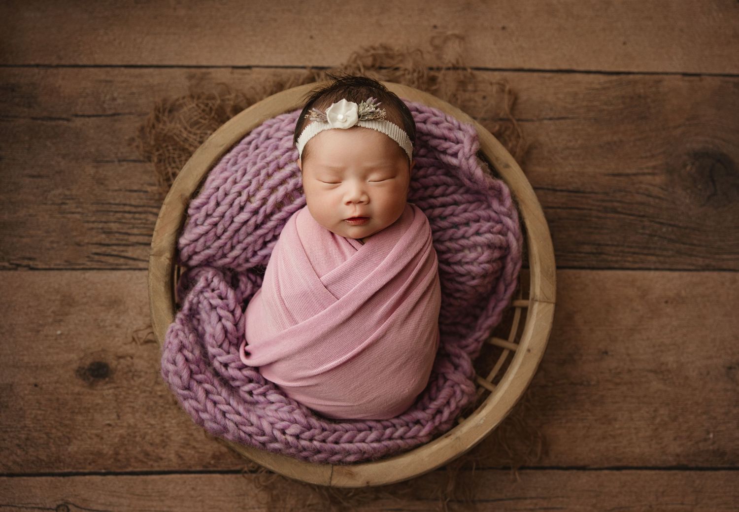Newborn baby sleeping peacefully in a wooden bowl with purple knit blanket on rustic wood floor.