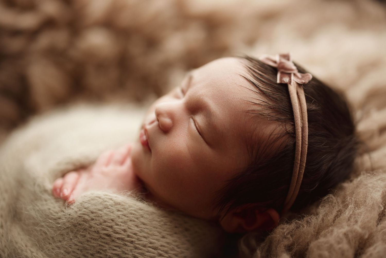 Peaceful newborn baby girl sleeping wrapped in knit blanket wearing pink bow headband, professional newborn photography.