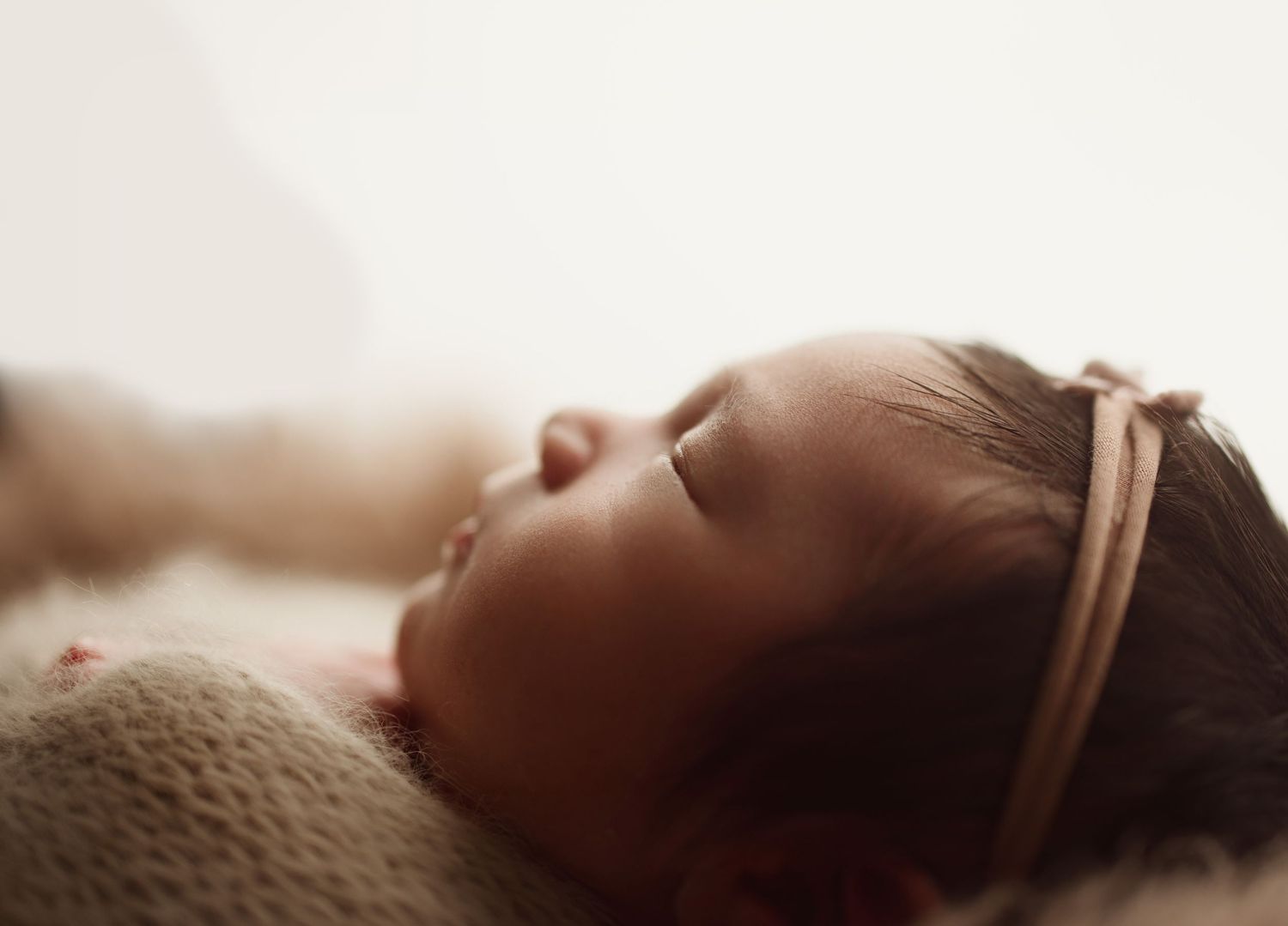 A peaceful newborn baby sleeping with eyes closed, wearing a delicate pink headband, wrapped in a soft cream blanket.