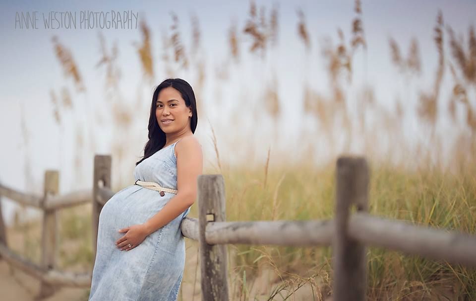 Pregnant woman in light blue dress leaning on a wooden fence near sea oats at a beach setting. va beach maternity photographer