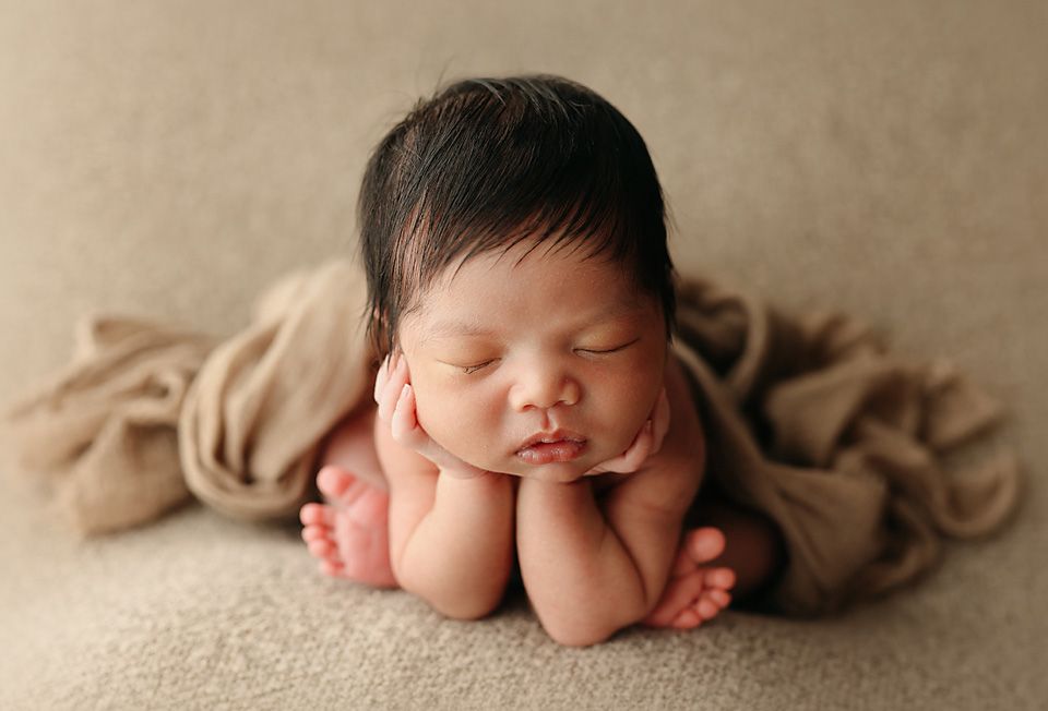 Peaceful sleeping newborn baby with dark hair posed with fists under chin on a beige blanket.