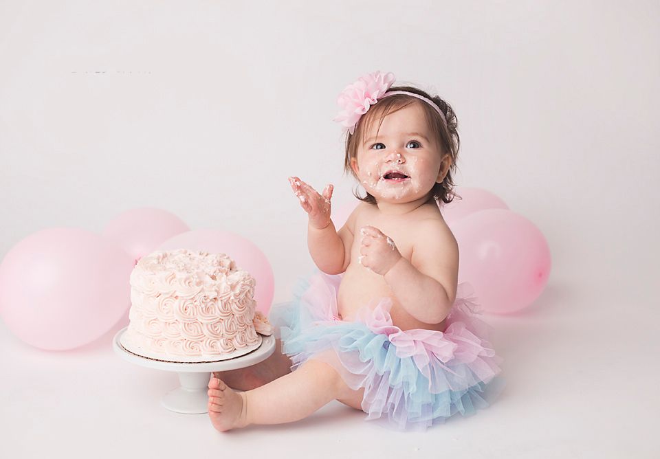 Happy baby girl in rainbow tutu celebrating first birthday with pink cake and balloons.