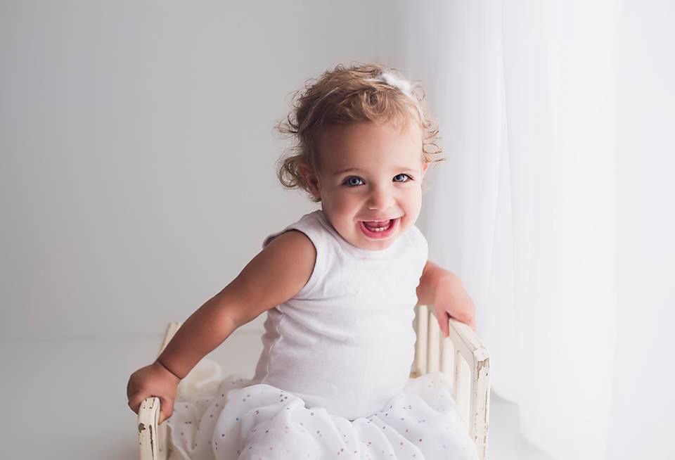 Happy toddler girl with blonde curls in white dress and tutu laughing inside a small white crib. baby photography Richmond