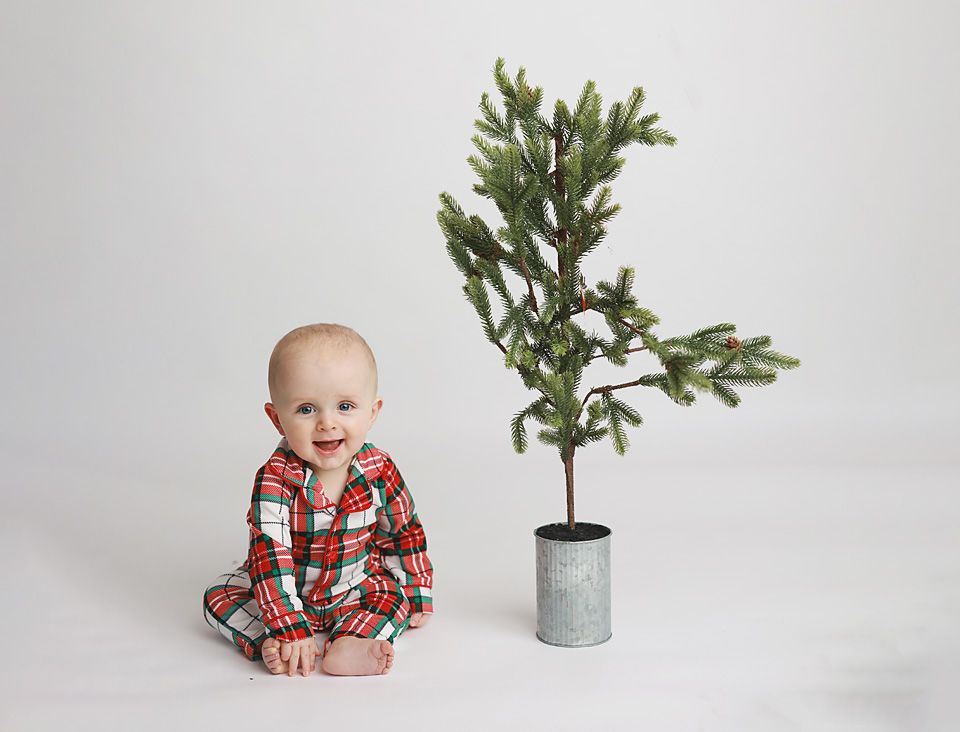 Happy baby in red plaid Christmas pajamas sits beside a small potted pine tree on white backdrop. studio photography baby Chesterfield