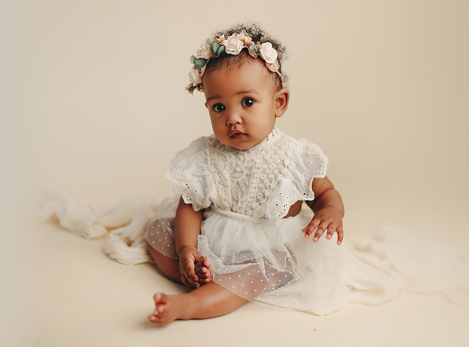 Adorable baby girl in white lace dress and floral crown sitting on cream background for professional portrait. baby photographer Richmond