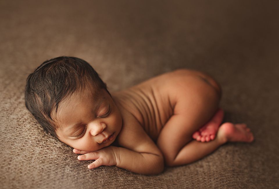 Sleeping dark-skinned newborn baby curled up peacefully on soft brown textured blanket.