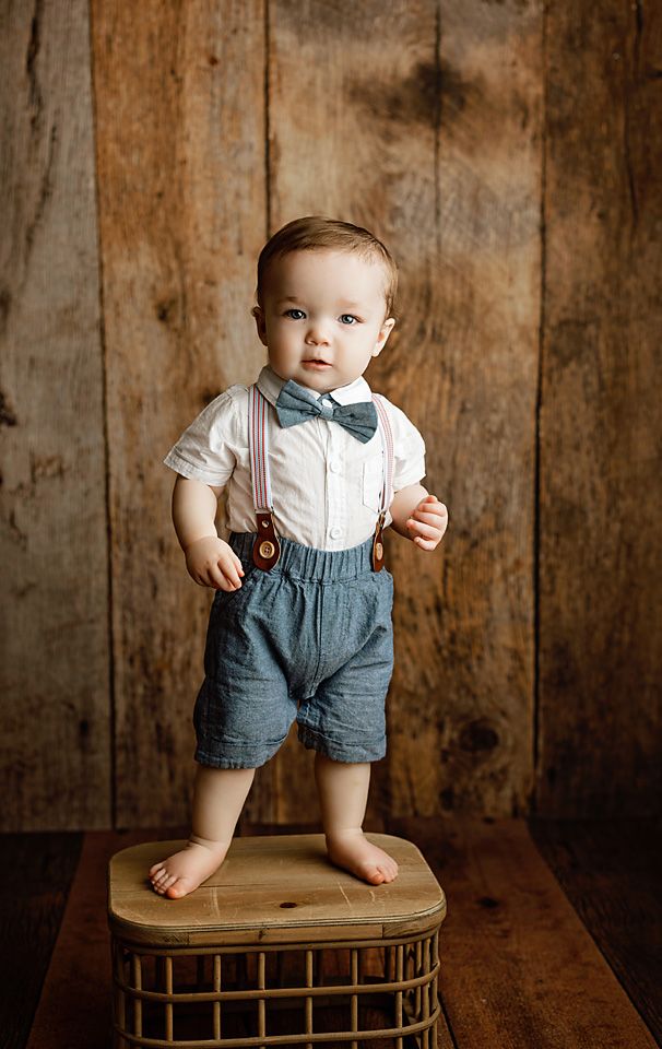 Cute toddler boy in suspenders and bow tie standing on wicker basket for birthday photos.