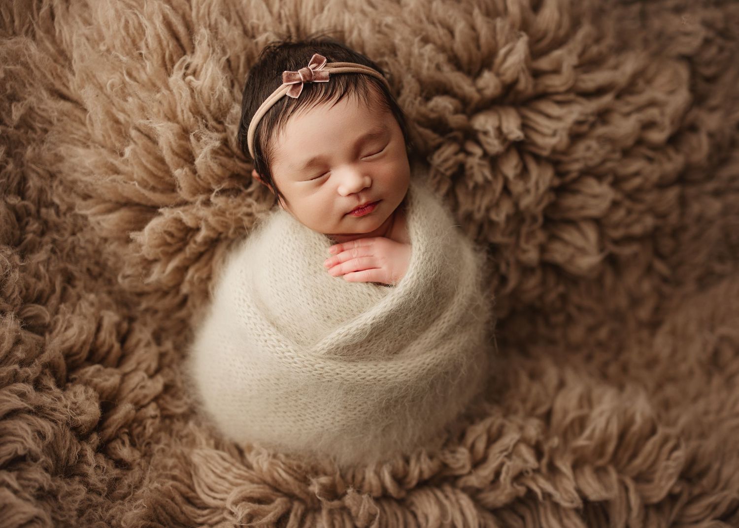 Newborn baby girl swaddled in cream wrap, sleeping peacefully on fluffy brown rug with floral headband.