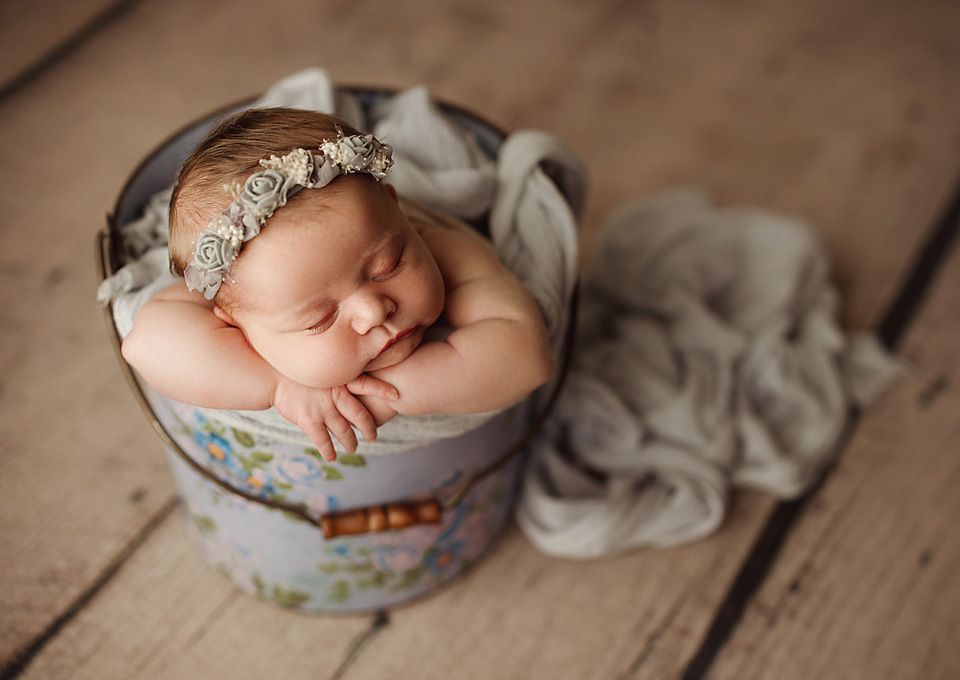 Sleeping newborn baby with floral headband curled up in decorative painted bucket on wood floor. Studio photography newborn Chesterfield