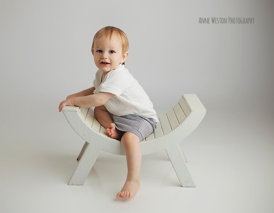 Cute toddler boy in white shirt and shorts sitting on a modern white wooden bench.