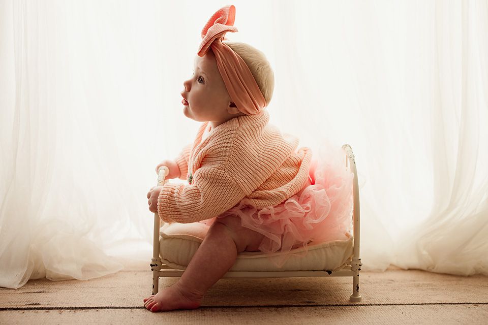 Baby girl in peach knit sweater and tutu sitting sideways on a miniature bed by sheer curtains.