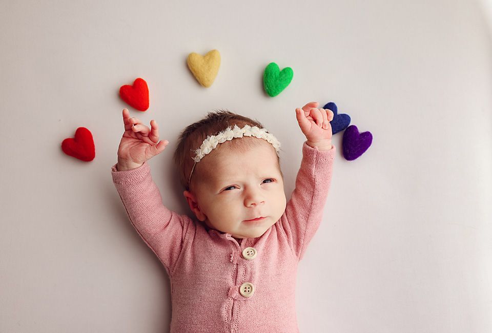 Baby girl in pink onesie lying on white background surrounded by colorful felt hearts with arms raised.